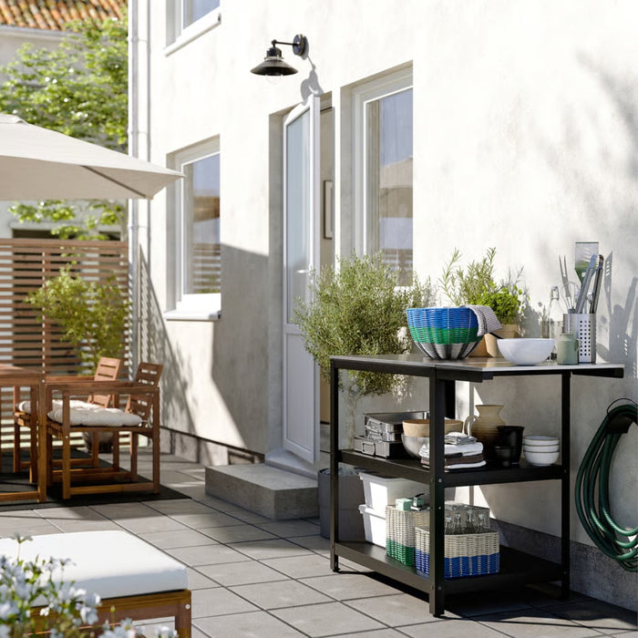 An inviting outdoor patio with wooden furniture and a large umbrella for shade. A black metal shelving unit against the house wall holds decorative baskets, bowls, potted plants, and utensils, enhancing the space with both functionality and charm. Greenery surrounds the area, creating a serene and relaxing environment.-10591422