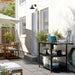 An inviting outdoor patio with wooden furniture and a large umbrella for shade. A black metal shelving unit against the house wall holds decorative baskets, bowls, potted plants, and utensils, enhancing the space with both functionality and charm. Greenery surrounds the area, creating a serene and relaxing environment.-10591422