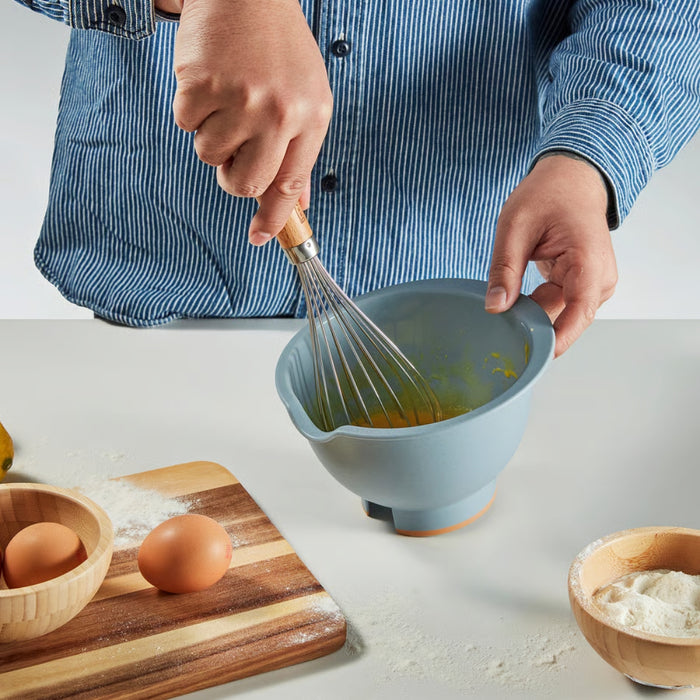 A person wearing a blue and white striped shirt using a whisk to mix ingredients in a blue mixing bowl. On the table, a wooden cutting board holds two eggs and a small wooden bowl with flour, with scattered flour adding a rustic kitchen feel.- 90607438