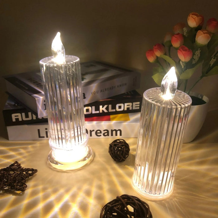 Two illuminated candle-like objects on a surface with books and flowers in the background.