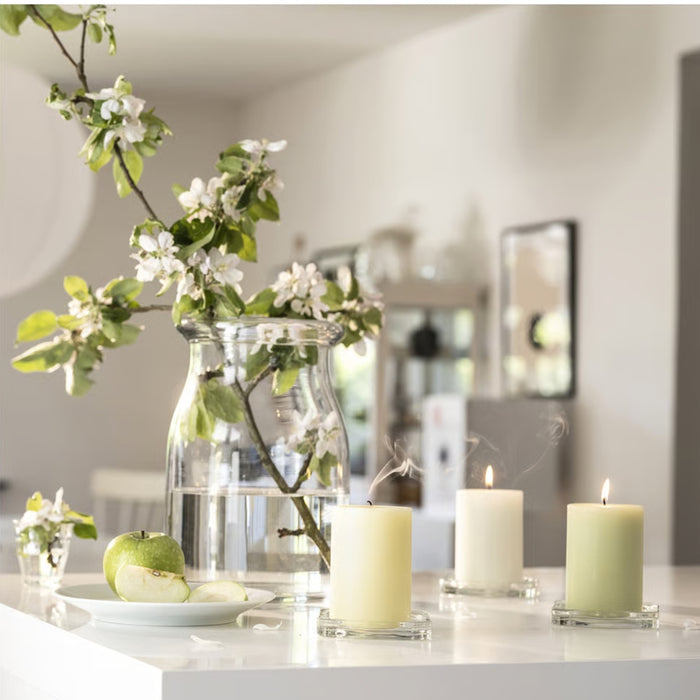 A cozy and elegant home setting featuring two lit IKEA LÖVSKOGSLUND Scented Pillar Candles with apple fragrance, placed on square glass coasters. Behind them, a large glass jar filled with water holds a branch with green leaves and delicate white flowers. The warm candlelight enhances the soothing and tranquil ambiance, making it a perfect addition to home décor-90582065
