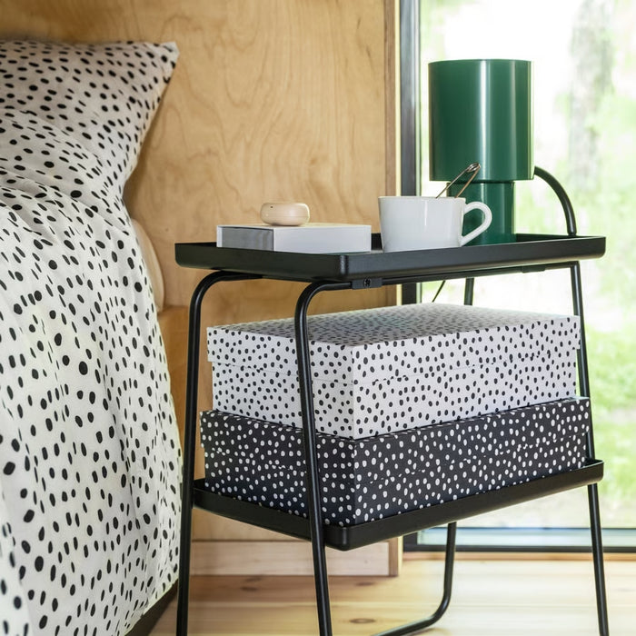 IKEA RÄTTAREN storage box set—two polka dot boxes (white with black dots and black with white dots) placed under a black metal bedside table. Table holds a white mug, book, wooden object, and green lamp. Bed beside it features matching polka dot bedding, with a wooden wall and window view of greenery in the background – 90597986.