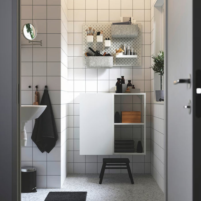 A modern bathroom with a white tiled wall featuring an IKEA SKÅDIS pegboard setup. The pegboard holds various toiletries, containers, and a small black towel. Below it, a white storage cabinet with open shelves houses folded towels, bottles, and a wooden box.-60599519