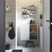 A modern bathroom with a white tiled wall featuring an IKEA SKÅDIS pegboard setup. The pegboard holds various toiletries, containers, and a small black towel. Below it, a white storage cabinet with open shelves houses folded towels, bottles, and a wooden box.-60599519