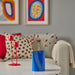 Blue vase with red flowers on a table in a living room with colorful artwork and cushions.