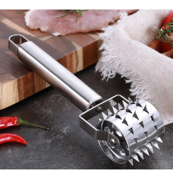 Metal meat tenderizer on a dark surface with red peppers and a wooden cutting board in the background.