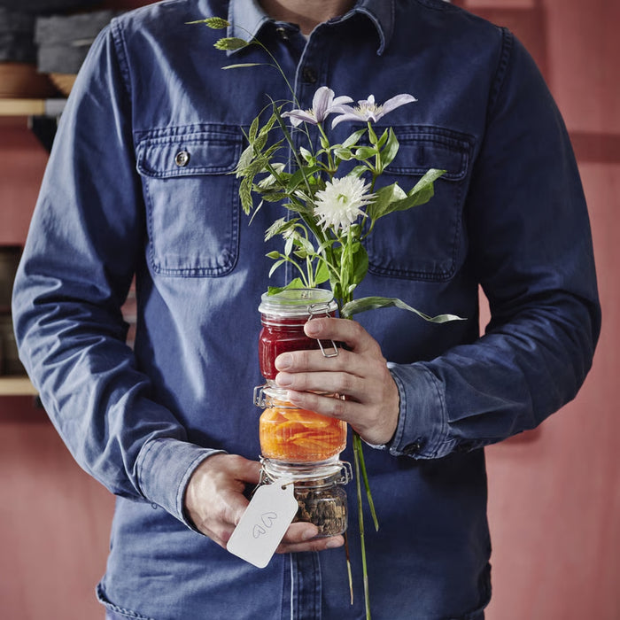 Person holding a stack of glass jars with flowers and a tag, wearing a blue shirt.