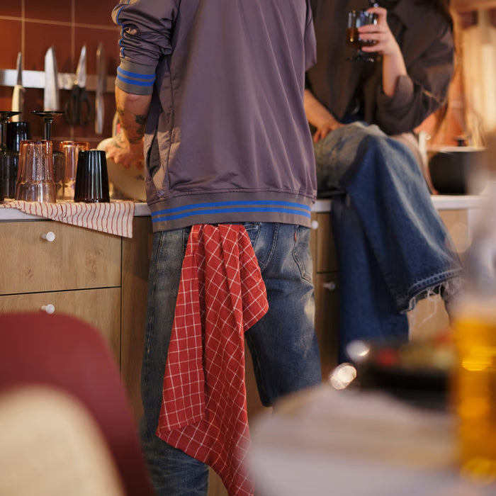 Person in a kitchen with a red checkered towel, another person sitting in the background.