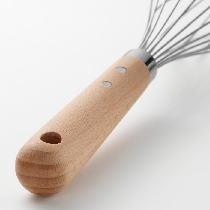 Close-up of the IKEA VARDAGEN balloon whisk lying flat on a wooden kitchen counter. The whisk’s natural beechwood handle features a smooth, ergonomic design with two metal rivets and a hanging loop at the end. The stainless steel wires form a classic balloon shape, ideal for whipping and mixing. The warm tones of the wooden surface and soft lighting emphasize the tool’s rustic charm and practical build -00581485.