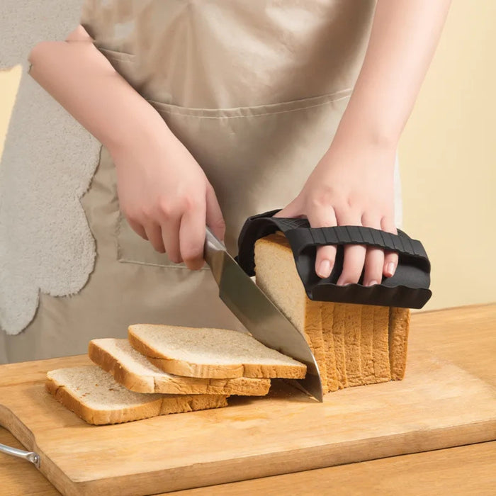 Person slicing a loaf of bread using a black bread slicer guide on a wooden cutting board. The guide ensures evenly sized slices, with three pieces already cut and neatly stacked. The individual is wearing an apron, and the setting suggests a clean, home kitchen environment—ideal for precision slicing and minimal waste.-1598452898401 13017586