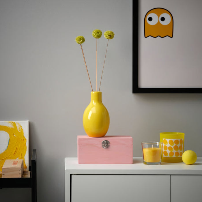 Yellow vase with floral arrangement on a pink box, glass with orange drink, and lemon on a white surface.