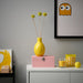 Yellow vase with floral arrangement on a pink box, glass with orange drink, and lemon on a white surface.