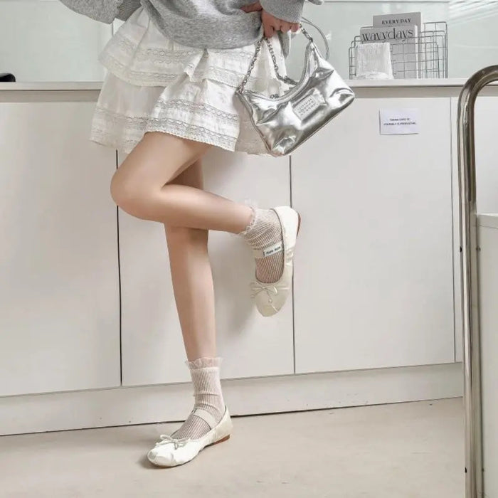 Person wearing a silver skirt, white shoes, and holding a silver bag in a minimalistic room.