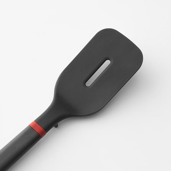 A black spatula with a rectangular head and a single elongated slot, attached to a dark gray handle with a red ring near the joint, displayed on a neutral background.