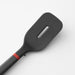 A black spatula with a rectangular head and a single elongated slot, attached to a dark gray handle with a red ring near the joint, displayed on a neutral background.