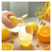 A hand squeezes a yellow citrus bottle and pours juice into a clear glass, surrounded by whole and sliced oranges on a wooden surface.
