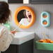 Child standing at sink with toothbrush, mouth open, facing circular orange mirror; colorful bathroom accessories visible.