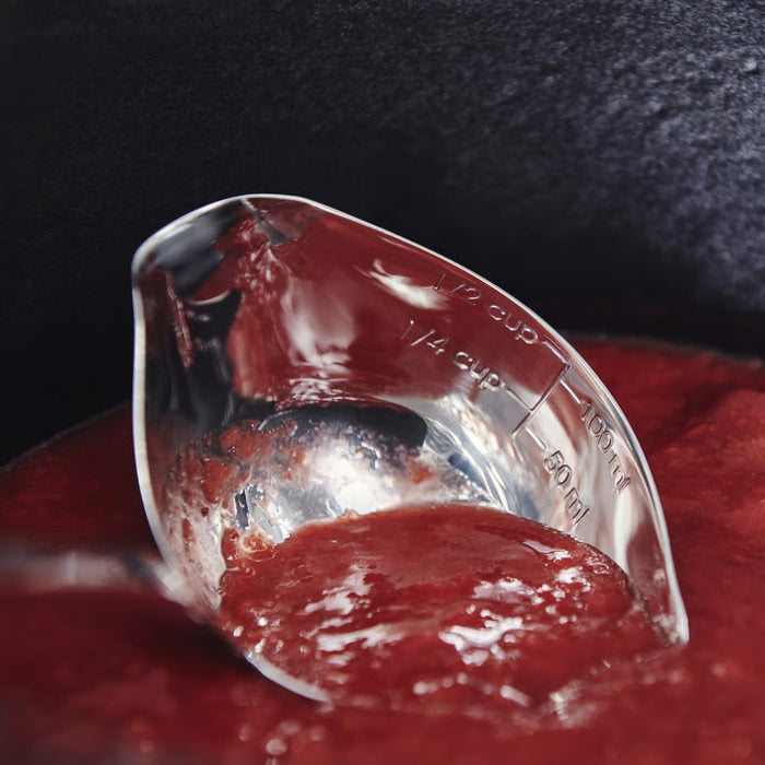 Transparent measuring cup with dual markings (cups and mL) partially submerged in thick tomato sauce inside dark cooking pot — ideal for precise liquid measurement in home cooking setups