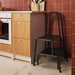 BÄSINGEN 2-step stool with handrail in anthracite, placed beside a wooden kitchen cabinet and oven. Red tiled wall and utensil setup highlight its utility in compact cooking spaces.– 30595754