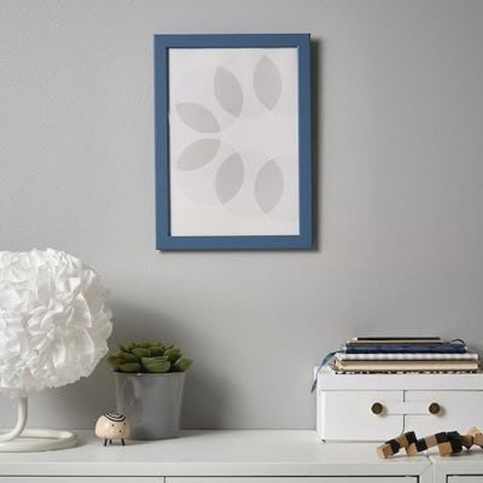 IKEA FISKBO frame in dark grey blue, 21x30 cm (8 ¼x11 ¾") placed on wall; displays split portrait of child’s photo and drawing – styled with white lamp, pen holder, toy car, and “HUGGET IS BEST” note for playful workspace decor - 20584930
