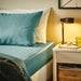Bed with teal bedding and pillows, nightstand with lamp and glasses, cozy bedroom setting.
