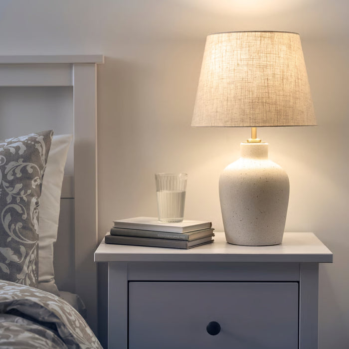IKEA BLIDVÄDER table lamp casting soft, warm light from its beige fabric shade, placed on a bedside table with books and a glass of water.

