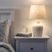 IKEA BLIDVÄDER table lamp casting soft, warm light from its beige fabric shade, placed on a bedside table with books and a glass of water.

