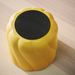 Yellow scalloped cylindrical container with flat black top, placed on a light wood surface.