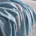 Close-up of the IKEA HOLMVI throw in soft blue—lightly woven texture with delicate white fringes along one edge, draped casually over a plush gray surface (possibly a sofa or bed). The gentle folds and fabric layering highlight its lightweight coziness and decorative appeal—ideal for adding warmth and visual softness to modern living spaces – 80604685.