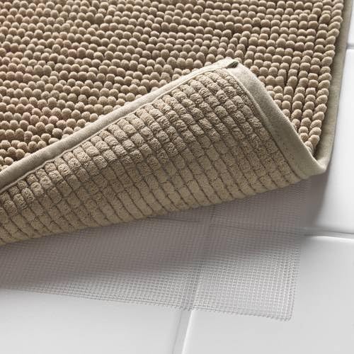Close-up of the IKEA TOFTBO pedestal mat in beige—corner of the mat is lifted to reveal its underside with a textured grid pattern. A white non-slip backing layer is visible underneath, positioned on a tiled bathroom floor, showcasing the mat's grip and safety features designed to prevent slipping –50610268