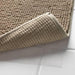 Close-up of the IKEA TOFTBO pedestal mat in beige—corner of the mat is lifted to reveal its underside with a textured grid pattern. A white non-slip backing layer is visible underneath, positioned on a tiled bathroom floor, showcasing the mat's grip and safety features designed to prevent slipping –50610268