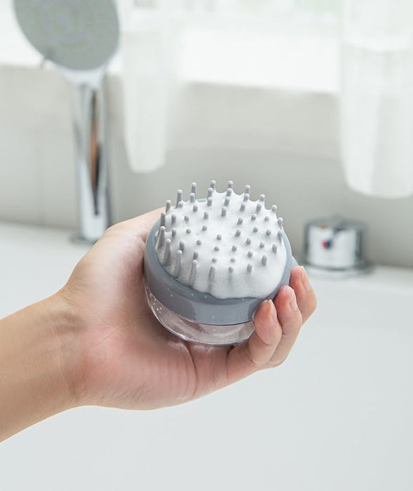 Gray silicone scalp massager with soft bristles, covered in foam, held in a hand for demonstration. Designed for shampoo application and gentle scalp massage to enhance cleansing and improve circulation. Showerhead and faucet in the background indicate use in a bathroom setting- 159845334888512362182