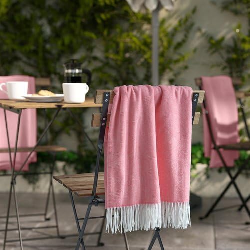 Outdoor dining setup with a soft pastel pink IKEA HOLMVI throw draped gracefully over the backrest of a wooden chair—chair features a pink fabric cover to match the throw’s hue. On the wooden table, white cups, a plate of food, and a black coffee pot are arranged neatly. The backdrop includes green foliage and a softly blurred umbrella, emphasizing a relaxed, stylish alfresco vibe – 00604689.