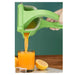 A green manual citrus juicer being squeezed by a hand, with orange juice pouring into a glass and lemon halves nearby on a tabletop.