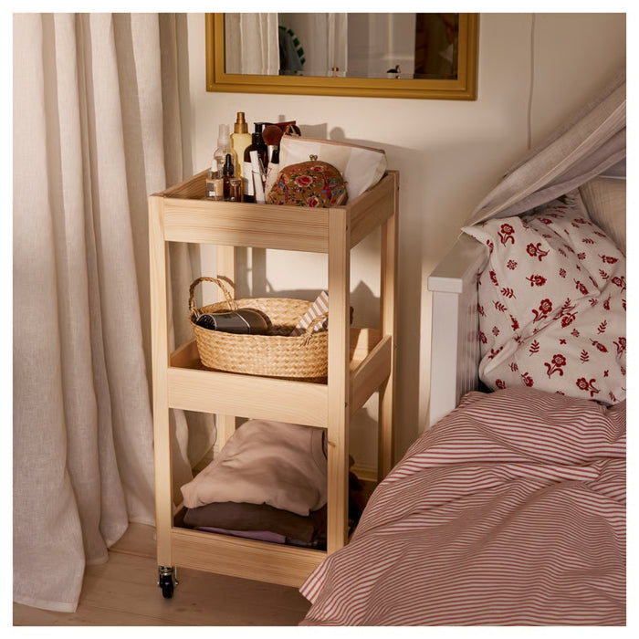  LOSHULT trolley beside bed in bedroom, top shelf with lotions and pouch, middle shelf with woven basket, bottom shelf with folded clothes, styled with striped bedding and floral pillows