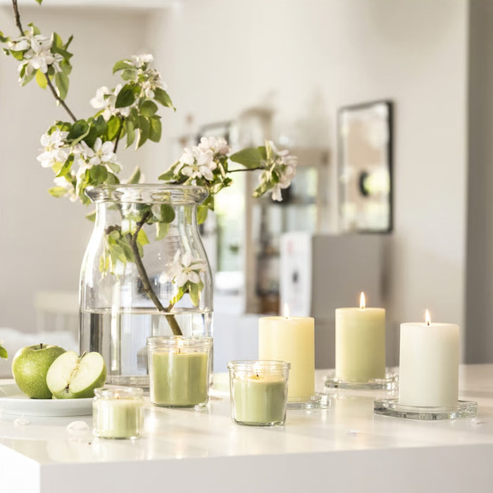 A stylish home decor setup featuring a transparent glass vase with water, holding a fresh branch with green leaves and delicate white flowers. Surrounding the vase are elegantly placed scented pillar candles in glass containers, emitting a soft, soothing glow. Nearby, a plate holds two green apples, one sliced in half, enhancing the fresh and inviting ambiance.-90582065