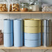 Stackable containers in various colors on a kitchen shelf with jars of snacks in the background.