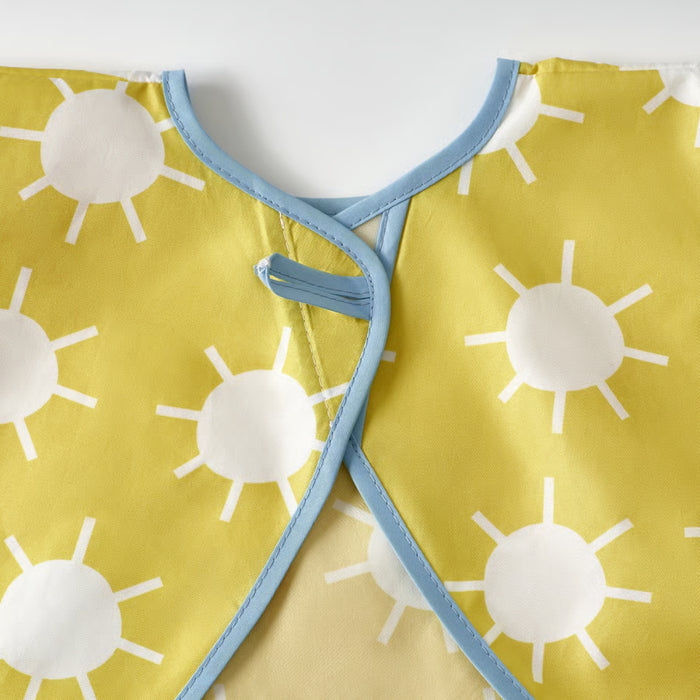 Close-up of IKEA KLADDIG bib in yellow/white; features cheerful white sun pattern on yellow fabric with blue trim and fastening loop – ideal for toddlers’ mealtime or craft protection - 00607145