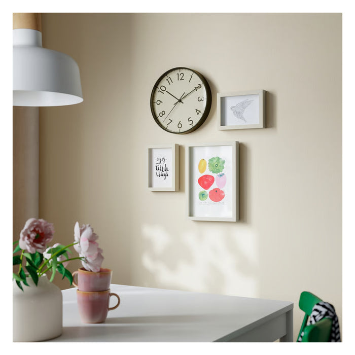 Wall with decorative clock and framed pictures, table with flowers and mugs in the foreground.