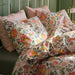 Neatly made bed with floral-patterned duvet cover and pillowcases in pink, yellow, green, and blue, paired with solid pink pillows and a gold bedside lamp.

