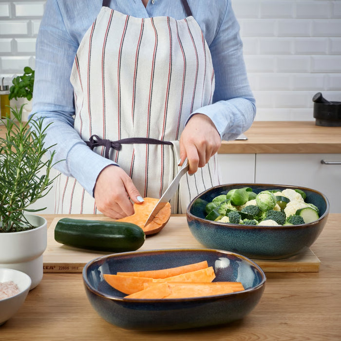 A cozy kitchen scene where a person in a striped apron is slicing sweet potatoes on a wooden countertop. Surrounding them are fresh vegetables including broccoli, cauliflower, zucchini, Brussels sprouts, and a bowl of sliced sweet potatoes. A potted rosemary plant and a small bowl of pink Himalayan salt complete the setup, with white subway tile backsplash and natural light enhancing the earthy, wholesome vibe -20531079