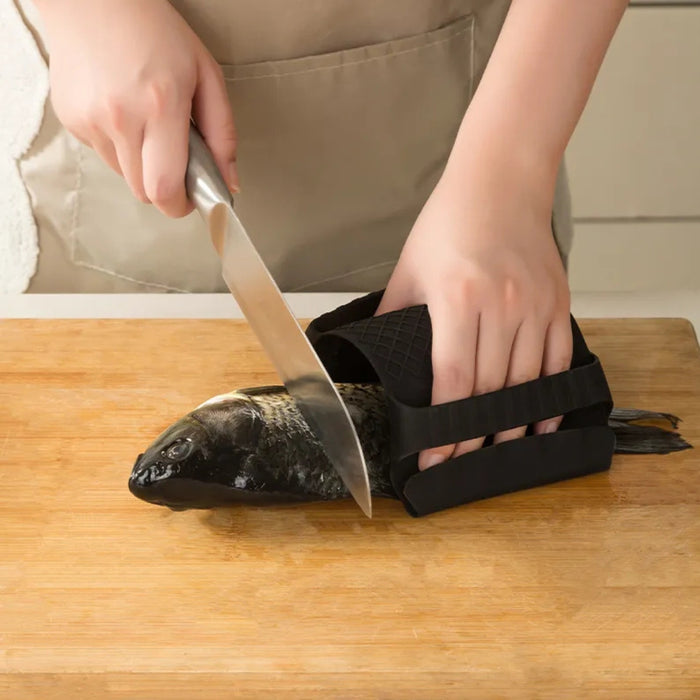 Person cutting a whole fish on a wooden cutting board using a knife, while securing the fish with a black silicone fish holder featuring a non-slip grip. The user is wearing a beige apron, and the scene is set in a kitchen environment—demonstrating safe, stable, and efficient fish preparation for cooking.- 1598452898401 13017586
