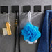 Five black wall hooks holding bath brush, necklace, pouf, and towel on gray wall