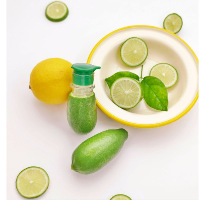 A small clear bottle with a green pump dispenser filled with green liquid, styled with whole and sliced limes and a lemon on a white background next to a yellow-rimmed bowl.