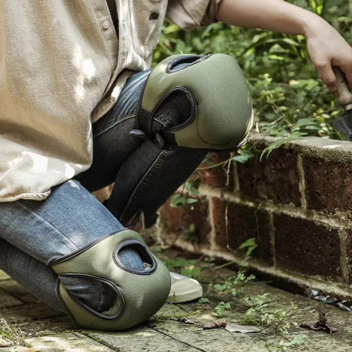 Person wearing green knee pads in an outdoor setting with a brick wall and greenery.