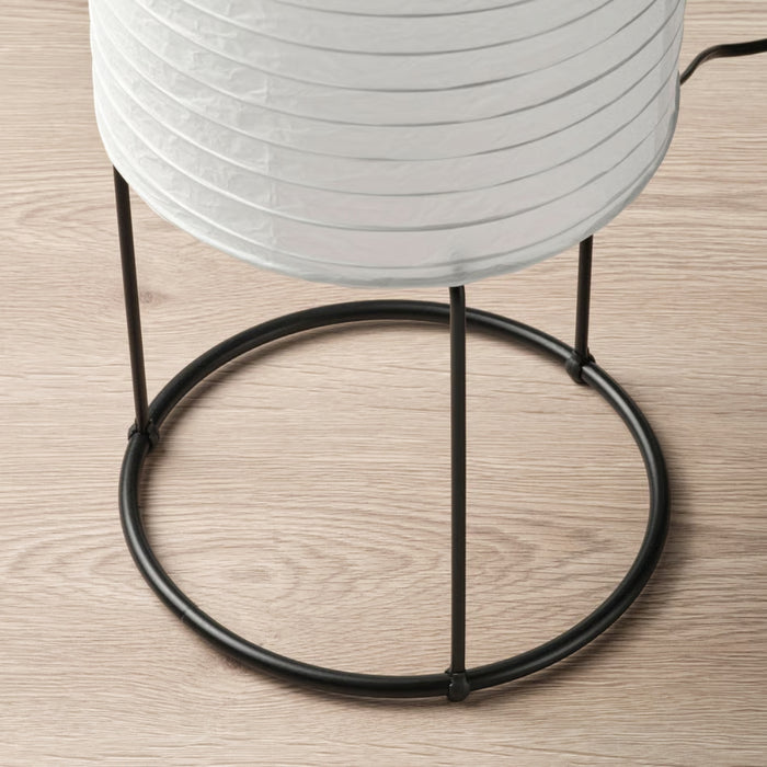 Close-up of IKEA STRANDAD floor lamp base with black metal frame and circular stand; white paper shade with horizontal ridges on wooden floor – 50594838