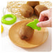 A hand uses a bright green fruit slicer to cut a kiwi on a light wood plate, with more kiwis and the green slicer visible in the background.