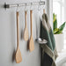  Four wooden cooking utensils and a stainless steel ladle hanging from hooks on a metal rod, with a striped towel and a potted plant in the background.