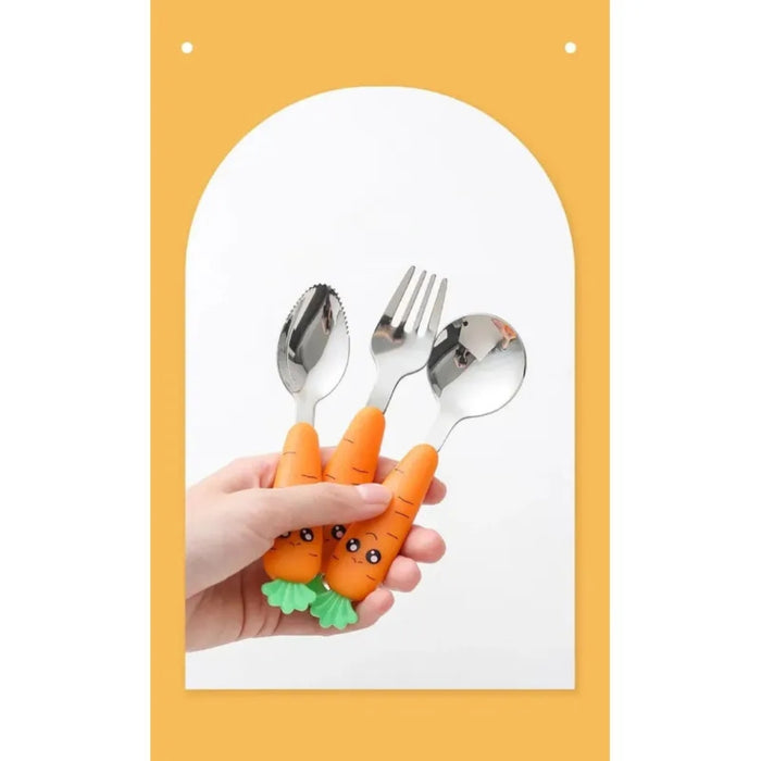 Hand holding a set of three carrot-themed children’s utensils—includes a fork, spoon, and serrated spoon with bright orange handles shaped like smiling carrots topped with leafy green ends; background features a light orange tone with a white arch, enhancing the playful and cheerful presentation – 1600002332714 12850097.