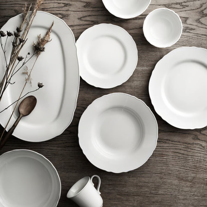IKEA UPPLAGA tableware collection, including an oval serving platter, plates, bowls, and a small cup, arranged on a wooden surface with a wooden spoon and dried plants—elegant white ceramic set for refined dining -60438022.